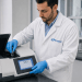 Lab scientist in a white lab coat and blue gloves inserts a sample into a benchtop analyzer in a modern laboratory, with a touchscreen display visible on the device showing data (546.1 nm).